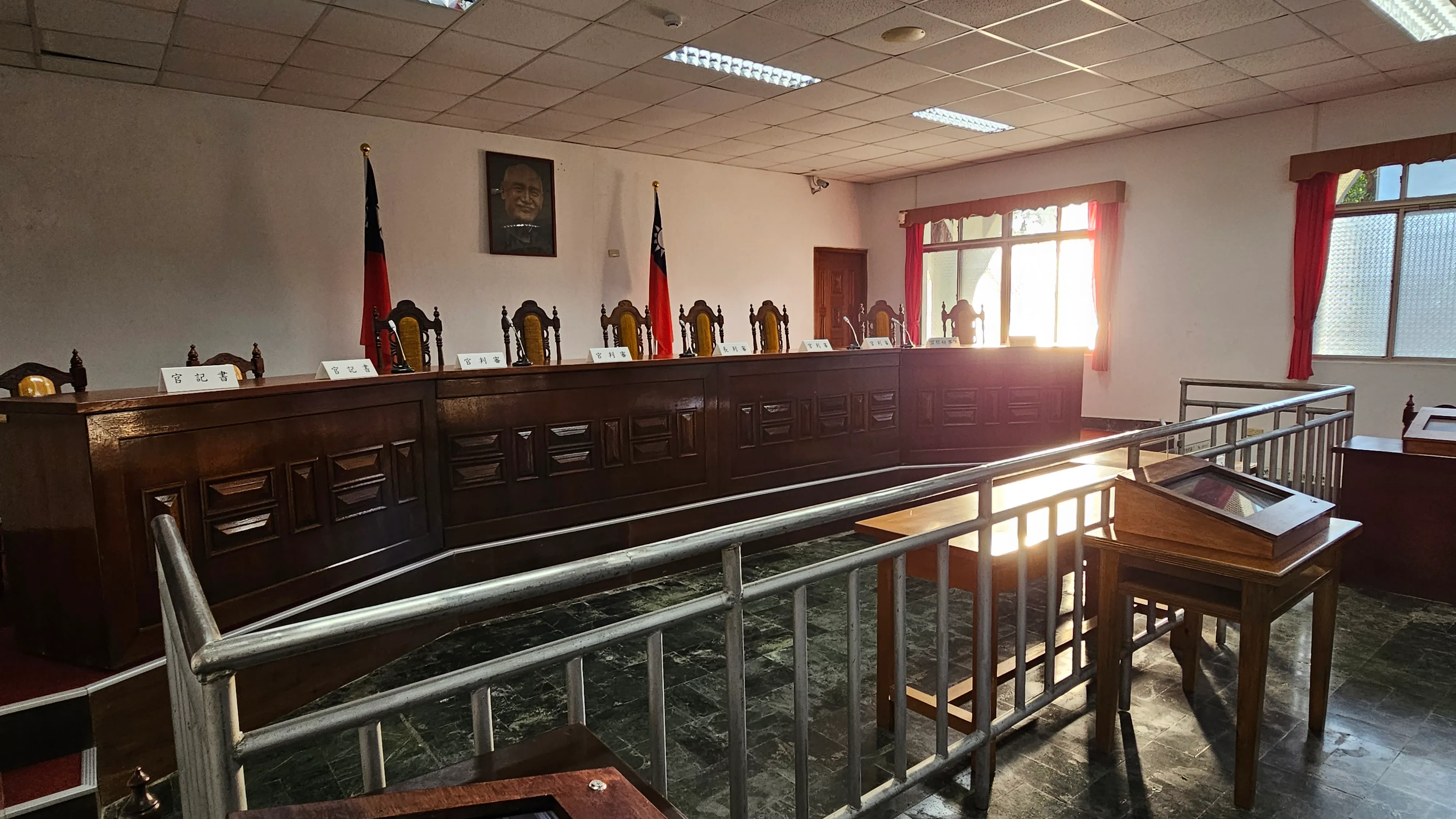 The restored First Court at Jingmei: a long dark wooden judges' bench with name plates and high-backed chairs, flanked by flags of the Republic of China, behind a metal defendants' rail