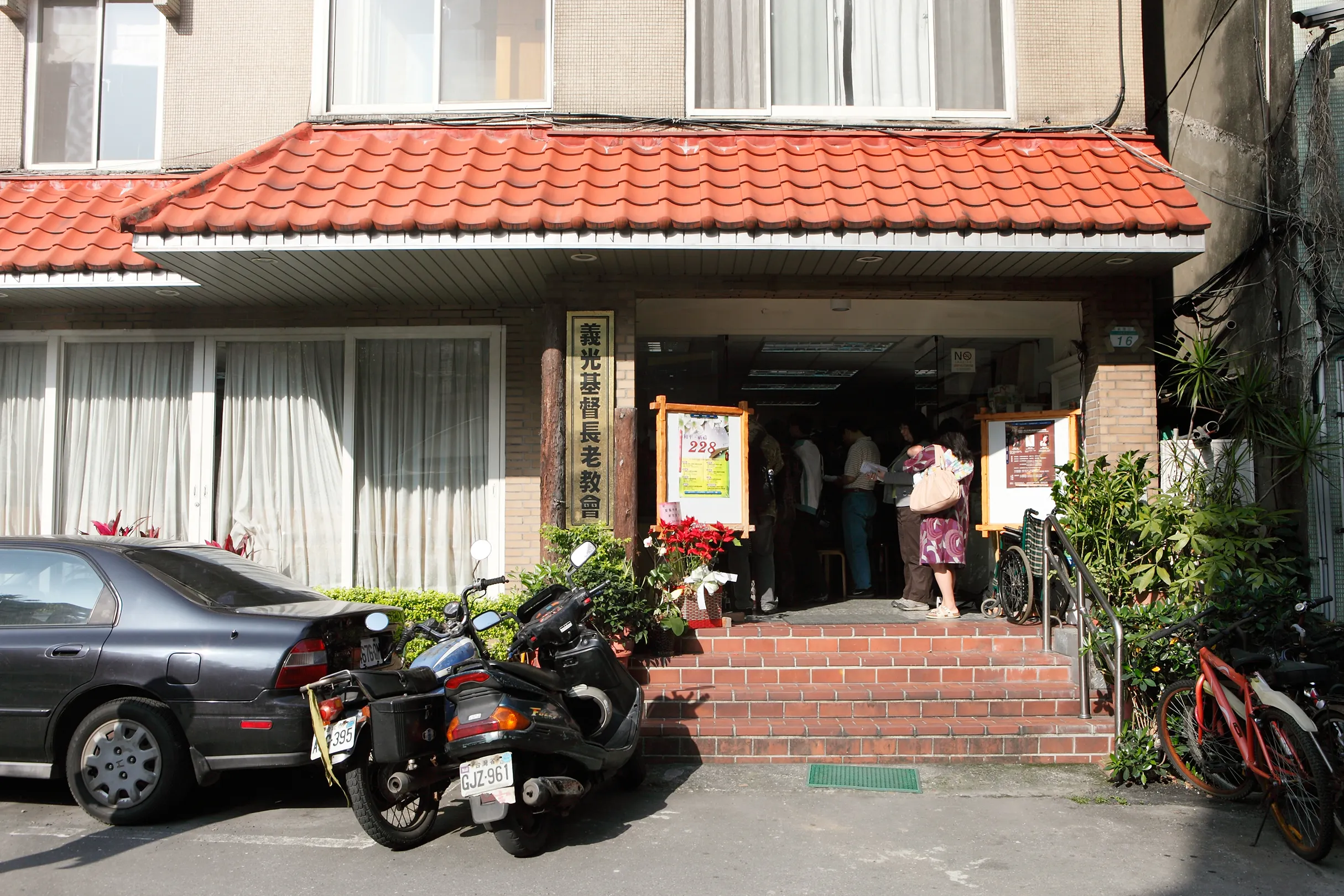 The front of Gikong Presbyterian Church: a modest brick row house with red-tiled eaves, a vertical sign reading 義光基督長老教會, and people gathered inside the open door at the top of red-brick steps