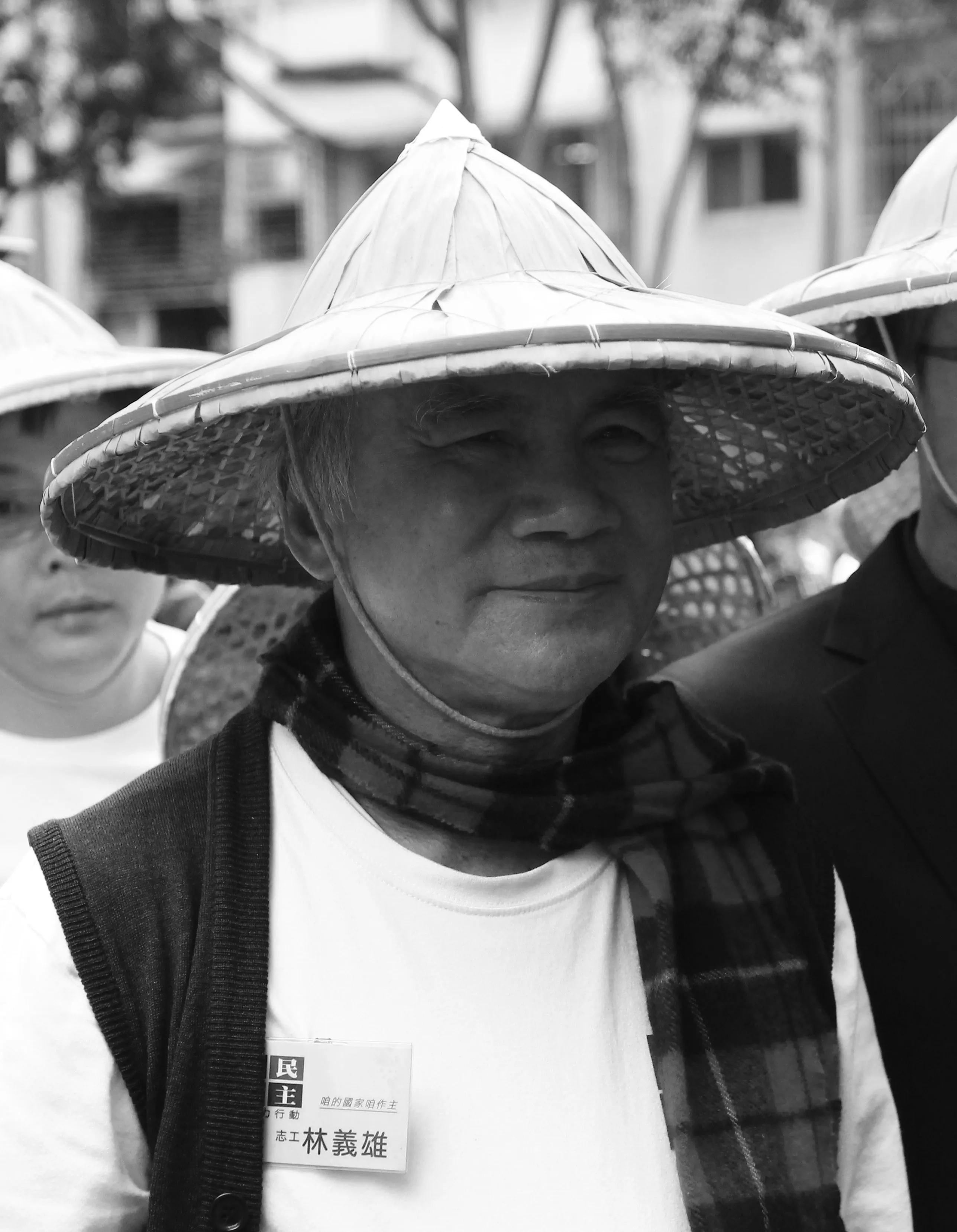 Black and white portrait of Lin Yi-hsiung in a woven bamboo farmer's hat and plaid scarf, faintly smiling, a volunteer name tag reading 林義雄 pinned to his shirt